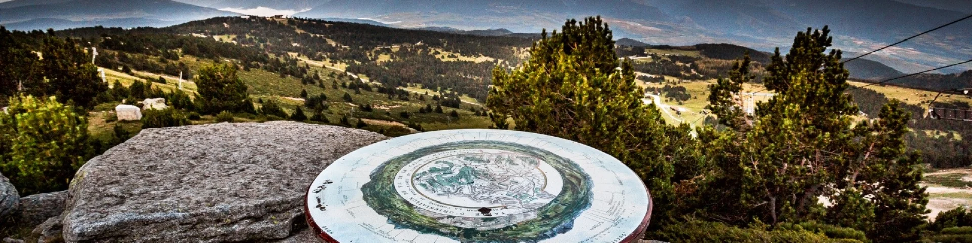 Table d’orientation en pierre sur un promontoire de montagne, offrant une vue panoramique sur les reliefs et vallées des Pyrénées-Orientales au coucher du soleil.
