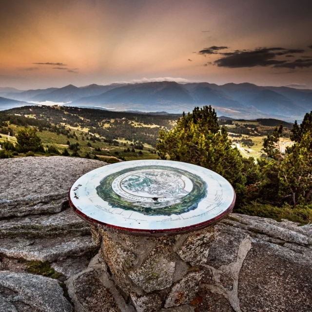 Table d’orientation en pierre sur un promontoire de montagne, offrant une vue panoramique sur les reliefs et vallées des Pyrénées-Orientales au coucher du soleil.