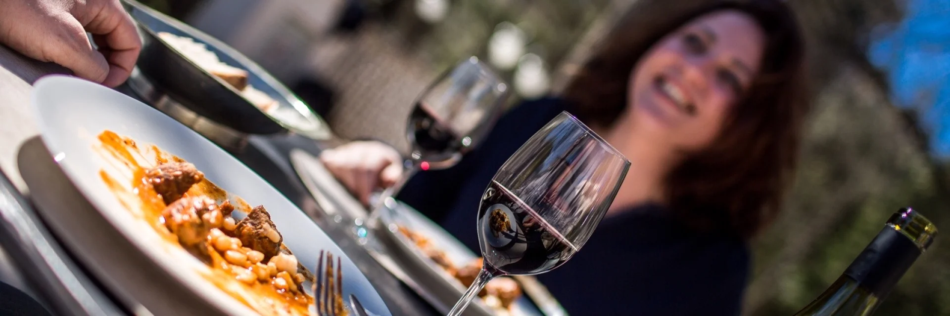 Repas convivial en terrasse avec deux personnes partageant un plat catalan et un verre de vin rouge sous le soleil.