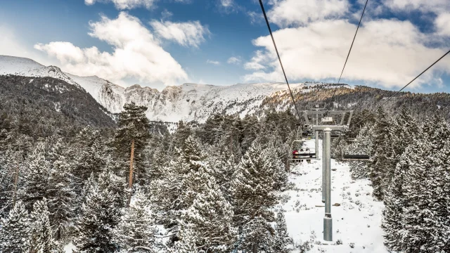 Vue depuis un télésiège survolant une forêt enneigée, avec les montagnes du Cambre d’Aze en arrière-plan sous un ciel partiellement nuageux.