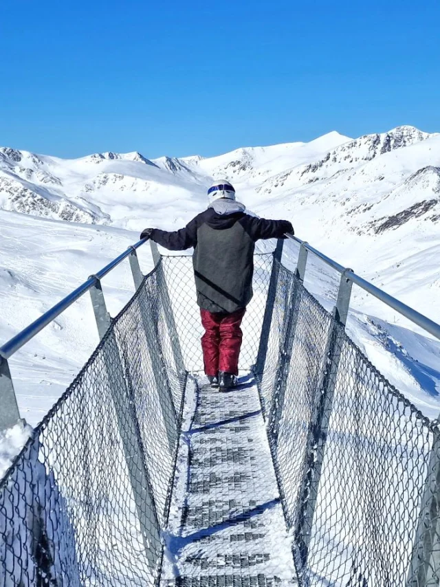 Personne debout sur la passerelle panoramique Le Pied dans le Vide à Porté-Puymorens, face aux montagnes enneigées sous un ciel bleu.