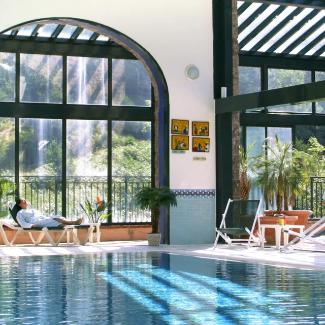Espace bien-être intérieur du Domaine de Falgos, avec piscine chauffée, baies vitrées ouvertes sur la nature et une cascade visible à l’extérieur. Deux personnes se détendent en peignoir au bord de l’eau.
