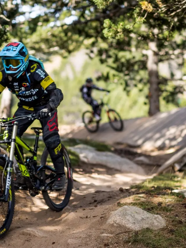 Deux personnes en VTT de descente roulant sur un sentier forestier avec passerelle en bois et terrain accidenté.