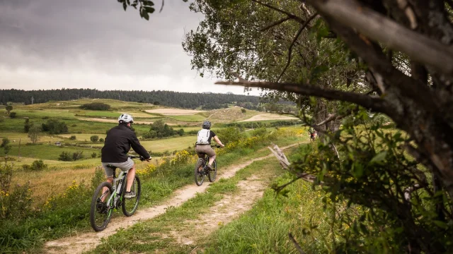 Deux personnes en VTT électrique roulant sur un sentier herbeux avec un paysage vallonné et verdoyant en arrière-plan.