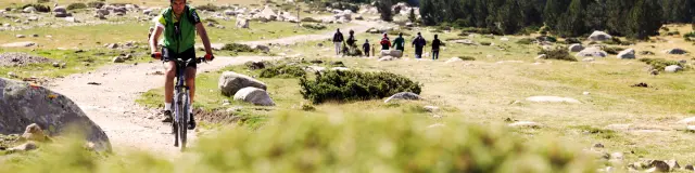 Cycliste en VTT avançant sur un sentier de montagne, avec un groupe de marcheurs au loin dans un paysage rocailleux.